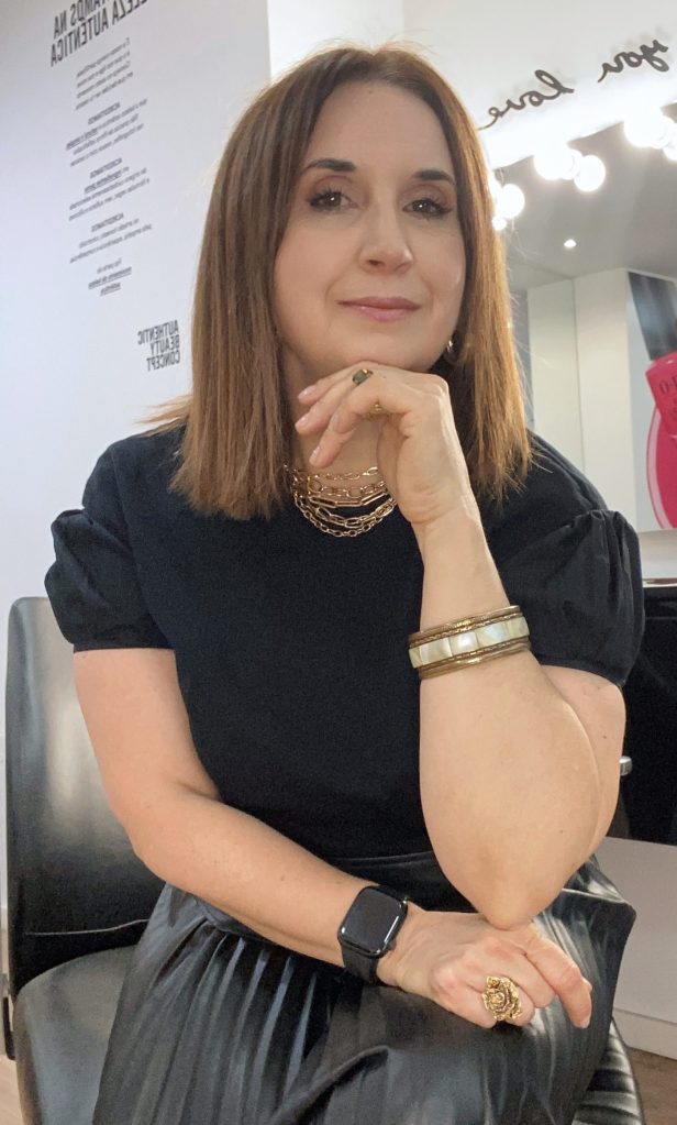 Mulher com cabelo castanho, usando uma blusa preta, sentada em uma cadeira. Ela está apoiando o queixo com a mão, sorrindo levemente. O fundo possui uma decoração moderna com espelhos e luzes.